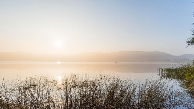 Ein nebliger Morgen an einem ruhigen See. Die Sonne geht langsam hinter den Bergen auf und spiegelt sich im Wasser.