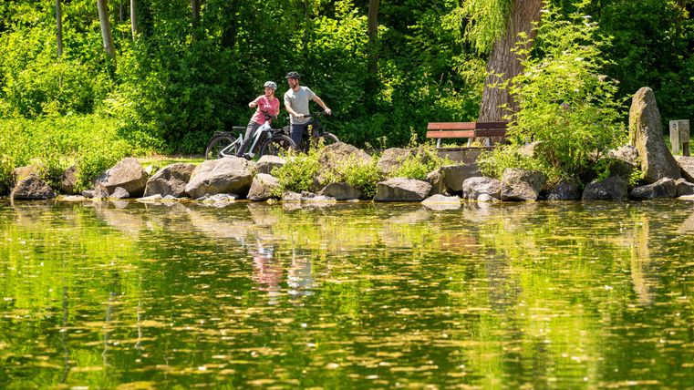 Ein Paar steht mit Fahrrädern am Ufer eines ruhigen Gewässers. Die Umgebung ist grün und üppig, mit reflektierenden Pflanzen im Wasser.