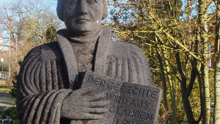 Eine Statue von Martin Luther, die ein Buch hält. Im Hintergrund sind Bäume und ein klarer Himmel zu sehen.