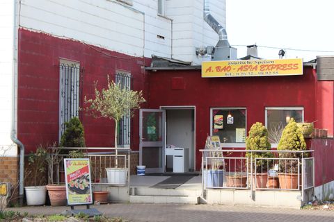 A red restaurant named "A. Ried - Asia Express" on the facade.Plants and an advertisement stand in front of the entrance.