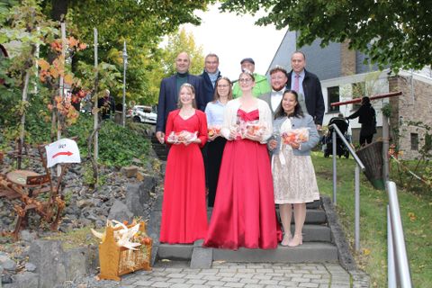 Een groep van zeven personen poseert vrolijk voor een lichte, met lampjes verlichte achtergrond. Ze dragen verschillende outfits en glimlachen naar de camera.