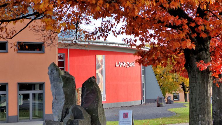 A modern building with a red facade, framed by colorful autumn trees. Informative stone sculptures stand in the foreground.