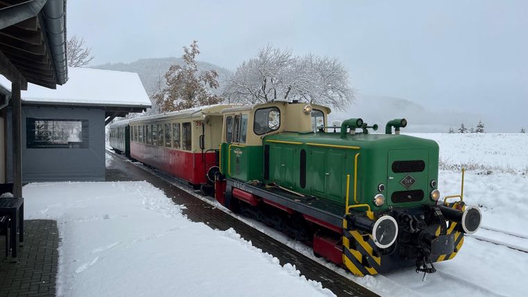 Eine grüne Lokomotive steht im Schnee an einem Bahnhof. Im Hintergrund sind schneebedeckte Landschaft und ein Zug zu sehen.