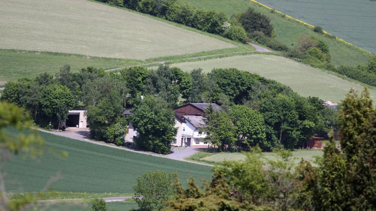 Ein ländliches Haus umgeben von Bäumen und Feldern. Die Landschaft ist grün und hügelig.