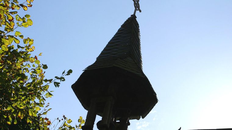 Ein historischer Kirchturm mit einem spitzen Dach und einem Kreuz an der Spitze. Der Himmel ist klar und blau, umgeben von Bäumen.