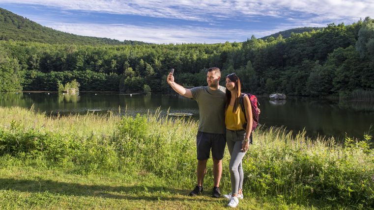 Ein Paar steht am Ufer eines Sees und macht ein Selfie. Im Hintergrund sind grüne Hügel und ein klarer Himmel zu sehen.