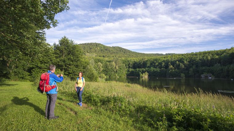 A hike in nature with two people by the shore of a body of water. In the background, trees and gentle hills can be seen.