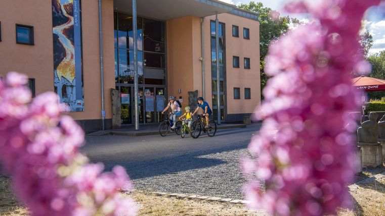 A modern building with colorful facades, surrounded by flowers. Some cyclists are passing by while the sun is shining.