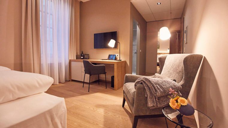 Ein modernes Zimmer mit einem bequemen Sessel und einem Arbeitsplatz. Die Wände sind in warmen Farben gehalten, und ein Fernseher ist an der Wand montiert.