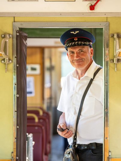 A train conductor stands in the door of a car and smiles. The interior is bright and the seating is clearly visible.