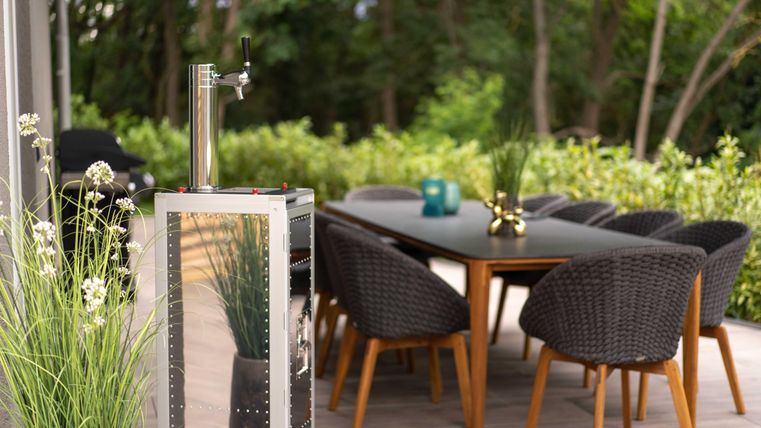 Ein stilvoller Außenbereich mit einem Tisch und Stühlen aus Rattan. Im Vordergrund steht ein Bierfass mit einer Zapfanlage.