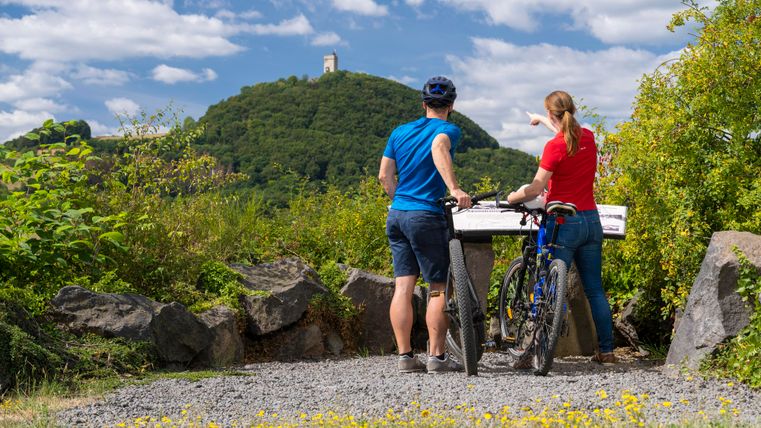 Zwei Radfahrer stehen an einem Aussichtspunkt und blicken auf einen Hügel mit einem Ausblick. Der Himmel ist blau und mit einigen Wölkchen verziert.