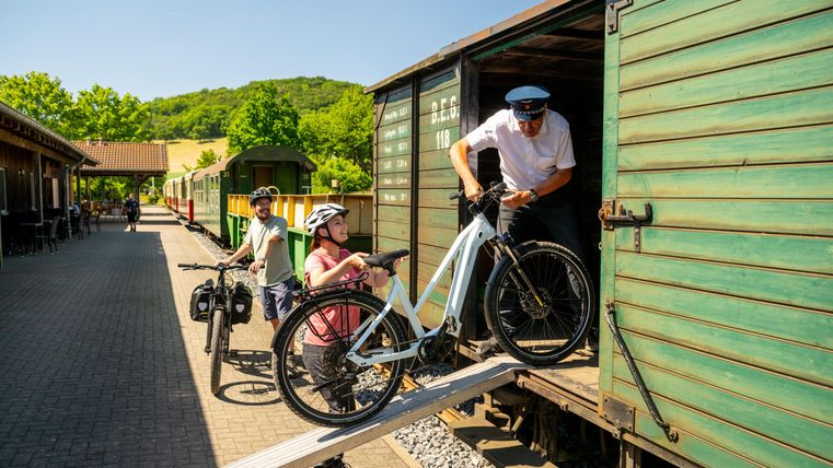 A train station with historical trains and cyclists. A train conductor is loading a bicycle into a carriage.
