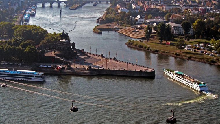 Eine malerische Flusslandschaft mit Schiffen und einer promontorierten Uferpromenade. Im Hintergrund sind Brücken und Häuser sichtbar.