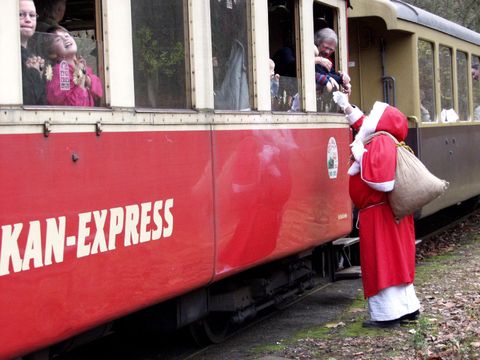Ein Weihnachtsmann steht vor einem Zug und verteilt Geschenke an Kinder, die aus den Fenstern schauen. Der Zug hat eine rote Farbe und heißt "Skan-Express".