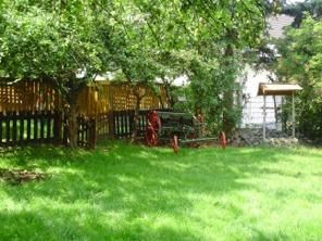 Ein grüner Garten mit einem alten, roten Wagen und einem Holzzaun. Im Hintergrund sind Bäume und ein Haus zu sehen.