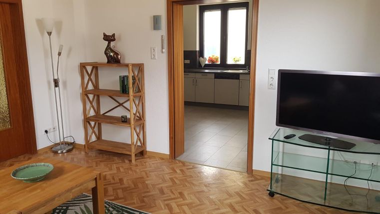 A cozy living space with a wooden floor and a shelf. In the background, the kitchen with windows can be seen.