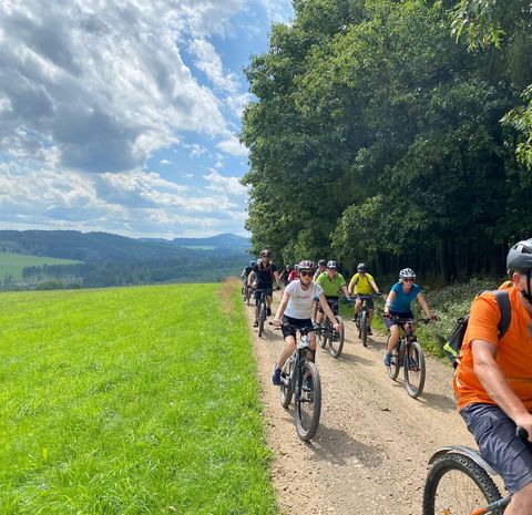 A group of cyclists is riding on a gravel path through a green landscape. In the background, trees and hills can be seen under a cloudy sky.