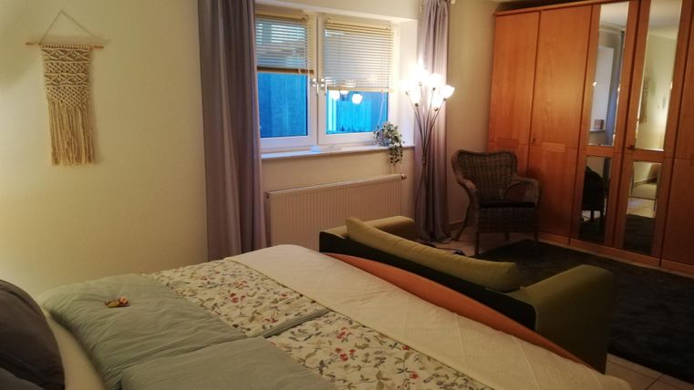 A cozy bedroom with two beds, an armchair, and a large wardrobe. The room has large windows with curtains and a beautiful plant.