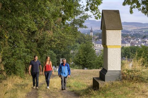 Three people are walking on a path with trees and a view of a city in the background. A tall, rectangular stone stands next to the path.