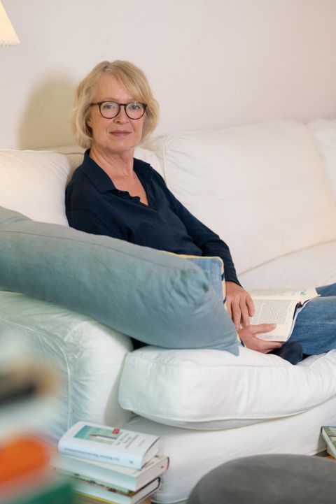 Een vrouw zit op een witte bank en leest een boek. Ze draagt een donkere blouse en heeft blond haar.