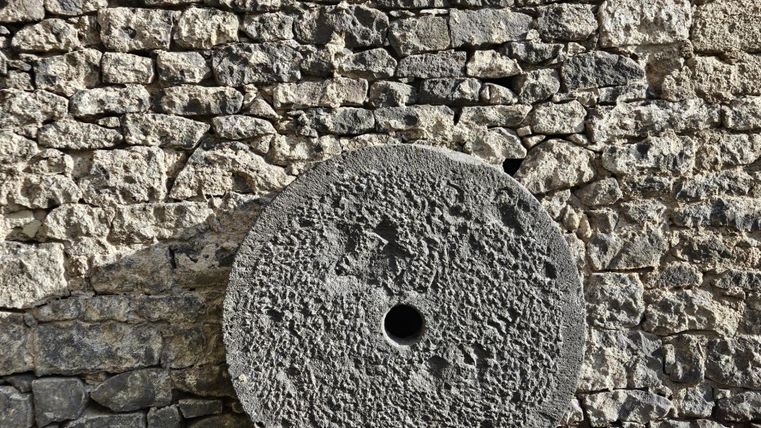 Ein großer, runder Mühlstein steht vor einer steinernen Wand. Die Textur des Steins ist rau und unregelmäßig.