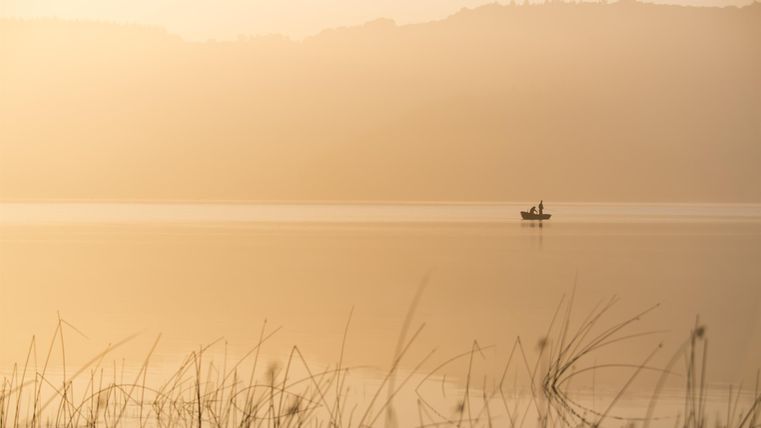 A peaceful lake with gentle hills in the background. A small boat floats on the water in the gentle morning mist.