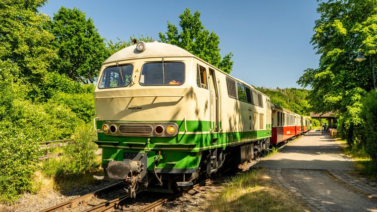 An old locomotive stands at a railway track in a green landscape. The train has several carriages in bright colors.