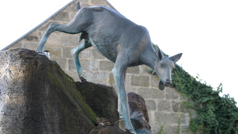 Eine Statue einer Ziege, die auf einem Stein steht. Im Hintergrund ist ein Gebäude und grüne Pflanzen zu sehen.
