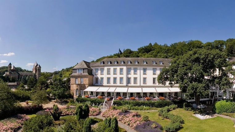 An elegant hotel surrounded by green trees and blooming gardens. The clear sky and beautiful architecture invite for a relaxing stay.