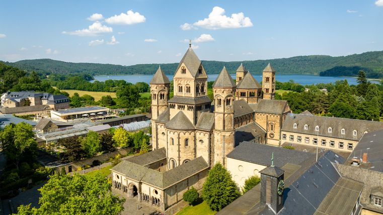 An impressive monastery complex with striking towers, surrounded by lush greenery. In the background, the lake and rolling hills are visible.