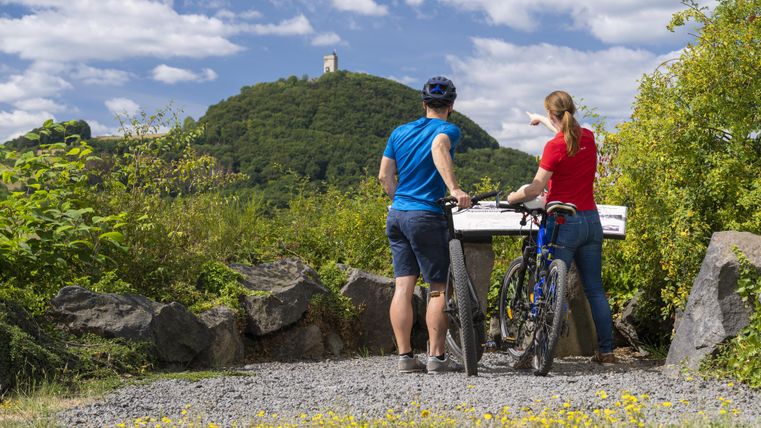 Ein Paar steht mit Fahrrädern vor einer Aussichtsplattform und blickt auf einen Hügel mit einem Turm. Der Himmel ist klar und es gibt viel grüne Natur um sie herum.