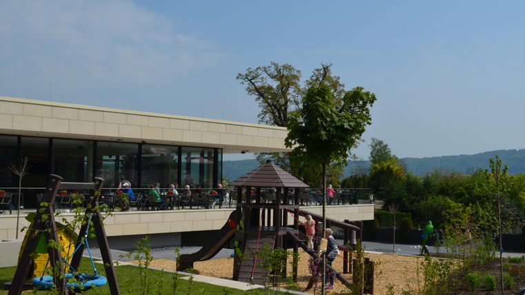 Ein moderner Spielplatz mit einer Rutsche und Sandfläche. Im Hintergrund ist ein Café mit Gästen und eine grüne Landschaft zu sehen.