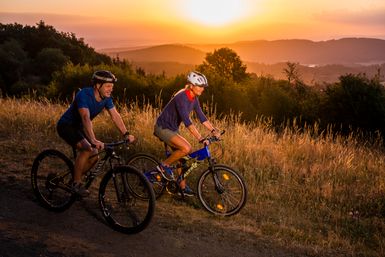 Zwei Fahrradfahrer auf Ihren Fahrrädern. Sie fahren einen Hügel hinunter. Im Hintergrund eine hügelige Landschaft im Sonnenuntergang.
