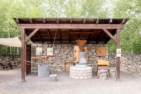 Ein überdachter Bereich mit Steinen und Werkzeugen. Es gibt Informationsschilder und Holzstrukturen, die auf eine historische Ausstellung hinweisen.
