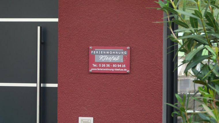 A sign reading "Holiday Apartment Klee" on a wall. The wall is red and there are green plants in the foreground.