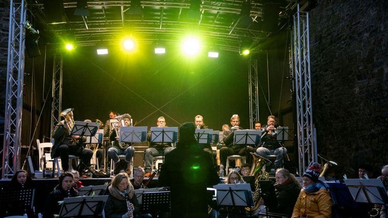Eine Musikkapelle spielt auf einer Bühne mit bunten Lichtern. Das Publikum sitzt davor und genießt die Darbietung.
