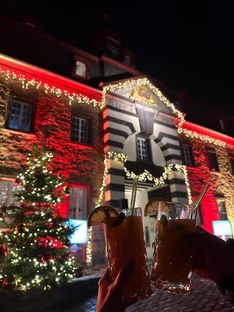 Ein festlich beleuchtetes Gebäude mit roten und weißen Lichtern. Im Vordergrund halten Personen zwei Cocktails mit Limettenscheiben.