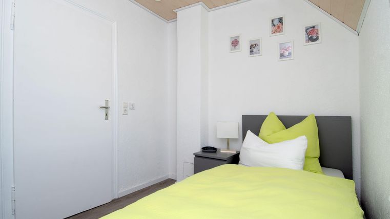 Ein kleines, helles Schlafzimmer mit einem Einzelbett und grüner Bettwäsche. An der Wand hängen Bilder, und der Raum hat ein modernes, minimalistisches Design.