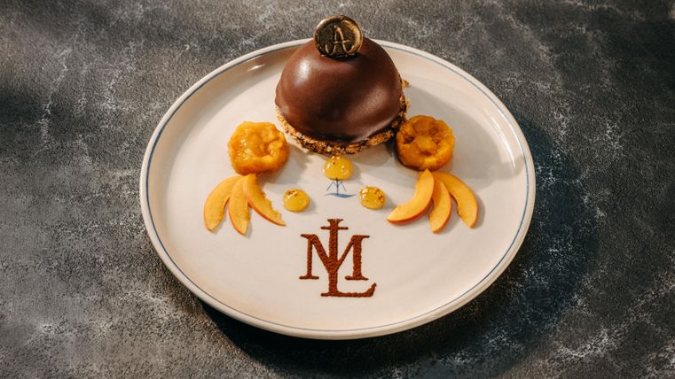 Ein kunstvoll angerichteter Teller mit einem Schokoladengebäck und dekorativem Obst. Das Design zeigt das Logo "ML" auf dem Teller.