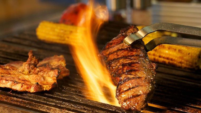 Ein saftiges Steak wird auf einem Grill zubereitet, während Flammen aufsteigen. Neben dem Steak liegen gegrillte Maiskolben und ein weiteres Stück Fleisch.