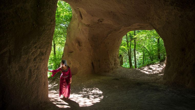 Eine Frau steht in einer Höhle, umgeben von Bäumen und natürlichem Licht. Die Szene strahlt eine ruhige, mystische Atmosphäre aus.