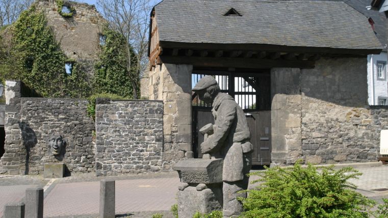 Eine Statue eines Handwerkers steht vor einer alten Mauer. Im Hintergrund sieht man ein Tor und Reste eines Gebäudes.