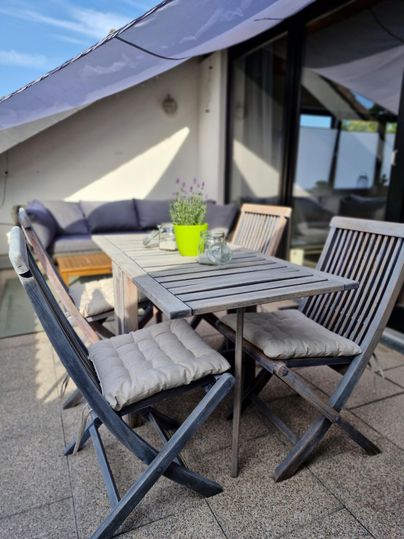 Ein gemütlicher Balkon mit einem Holztisch und vier Stühlen. Auf dem Tisch steht eine grüne Blume und es gibt ein paar Gläser.