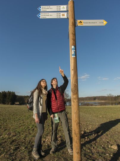 Zwei Wanderer stehen neben einem Wegweiser auf einem Feld. Der Himmel ist klar und blau, und sie zeigen auf die ausgeschilderten Wanderwege.
