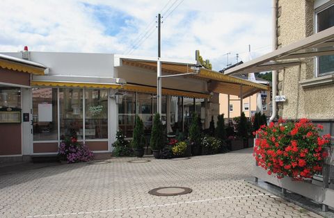 A small restaurant with large windows and yellow awnings. Surrounding the entrance area are plants and colorful flowers.