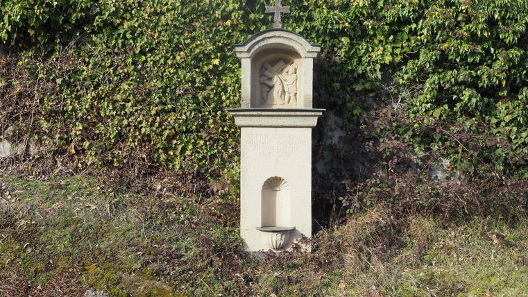 Eine kleine Kapelle mit einer Statue in der Nische steht vor einer grünen Hecke. Die Umgebung ist mit trockenem Gras und Erde bedeckt.
