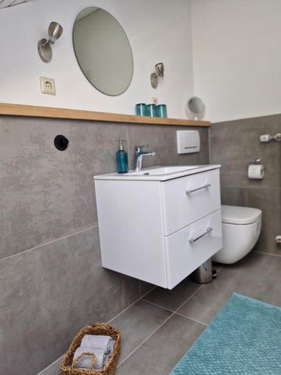 Ein modernes Badezimmer mit einem weißen Waschbecken und einem großen Spiegel. Der Fußboden ist grau gefliest, und ein türkisfarbener Teppich liegt vor dem Waschbecken.