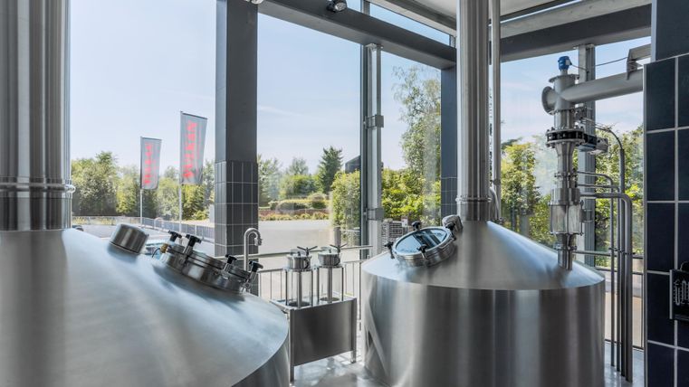 Eine moderne Brauerei mit glänzenden Edelstahlbehältern. Durch große Fenster ist viel Tageslicht und eine grüne Umgebung sichtbar.