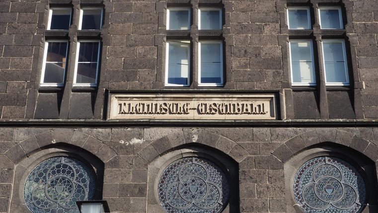 Ein historisches Gebäude mit einem dunklen Steinfassade und großen Fenstern. Über einem der Fenster steht der Schriftzug „Ehemals - Gasthaus“.
